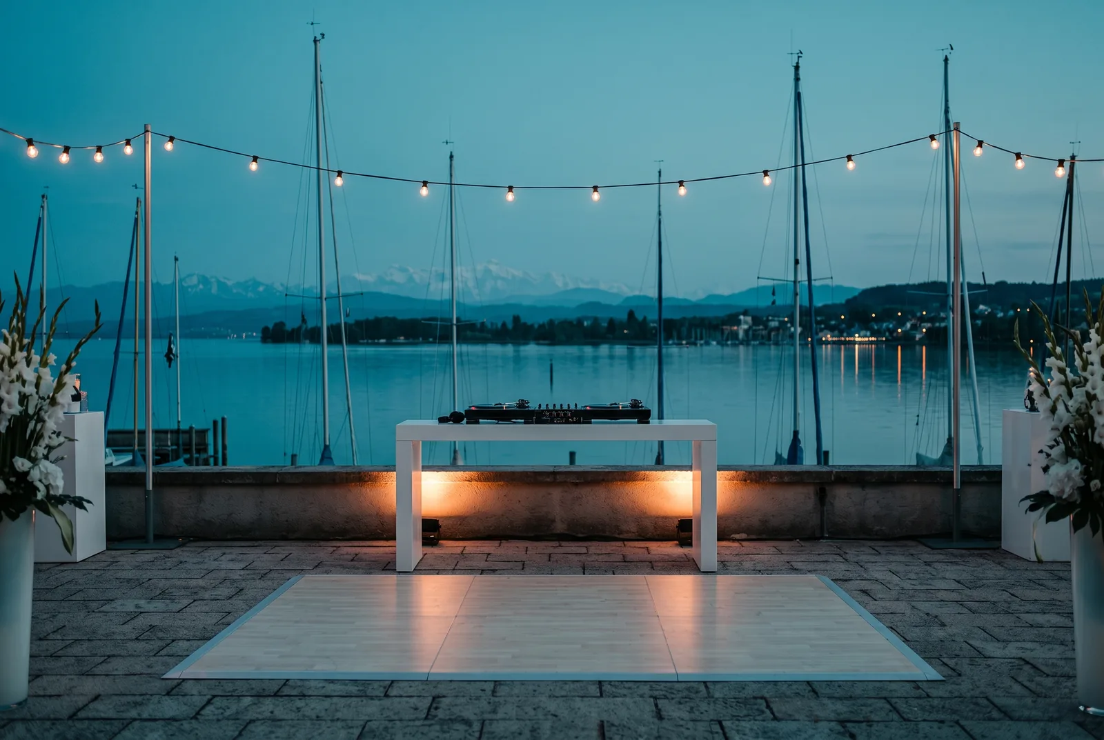 KI-Illustration eines DJ-Pults auf einer Seeterrasse am Bodensee zur blauen Stunde